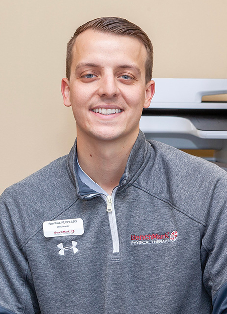 A person in a gray jacket with a name tag, smiling, in front of office equipment.